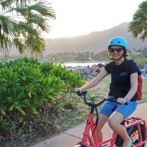 Person with blue helmet rides red bike on path through tropical scenery.