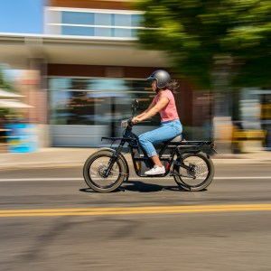 Person wearing helmet rides e-bike on city street, buildings blurred in background.